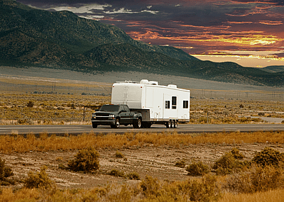 A black pickup truck towing a white camper trailer down a long highway.