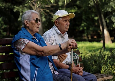 An older couple sitting on a bench in a park. An older woman wearing sunglasses, resting her hands on the cane.