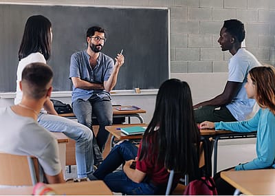 A male teacher sitting on top of the desk looking at the African American male student to his left in a classroom. The rest of the students are facing the teacher.
