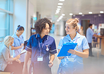 Two nurses walking down a hospital hallway while reviewing notes on a clipboard.