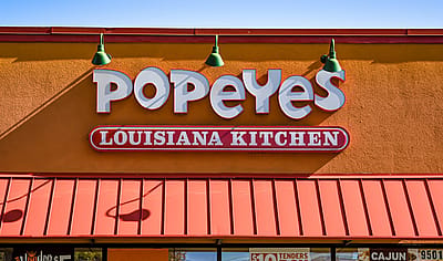 orizontal, medium closeup of "Popeyes" Louisiana Kitchen exterior facade brand and logo signage in white letters on a bright sunny day.