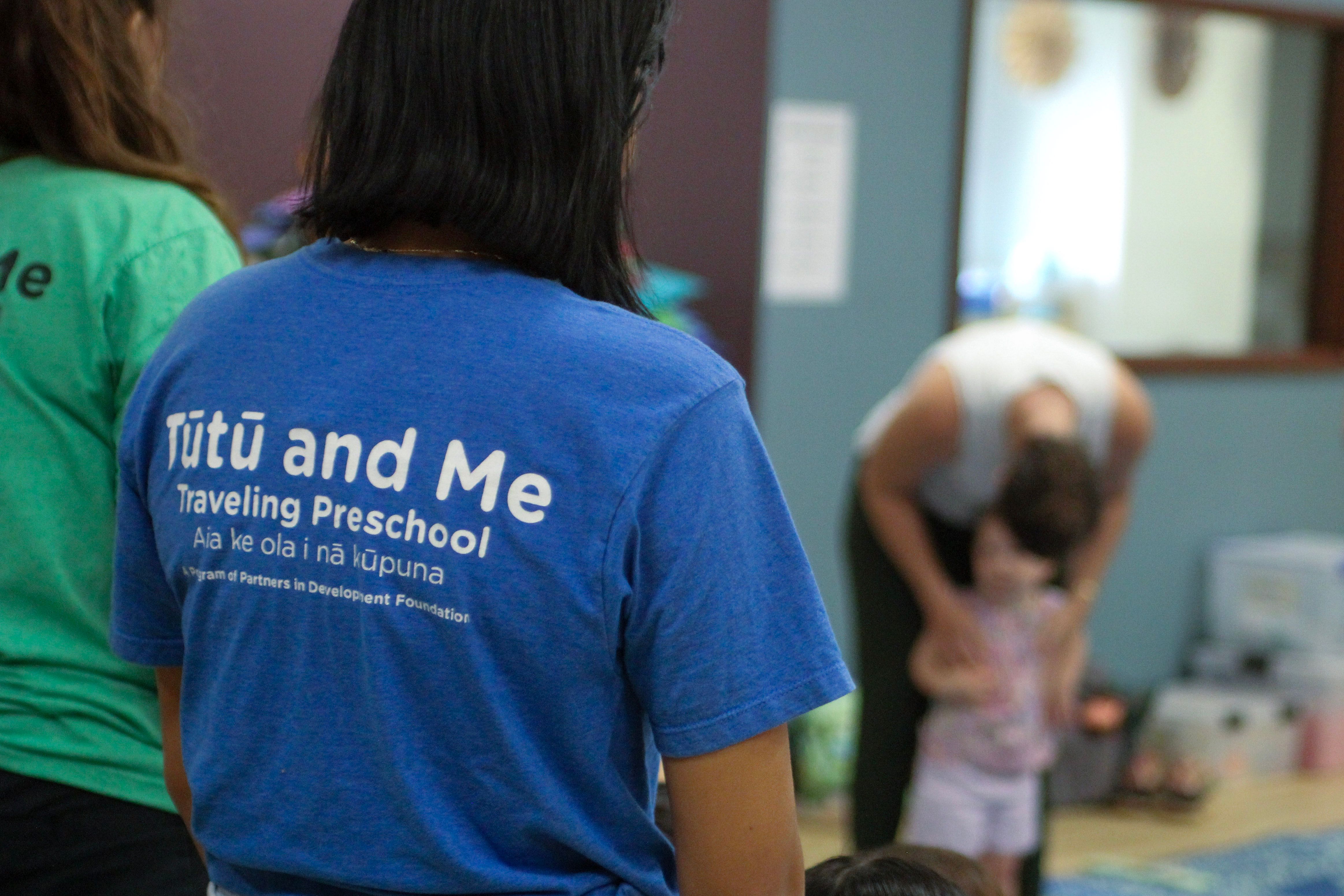 A person with a Tūtū and Me T-shirt looks at an adult interacting with a child.
