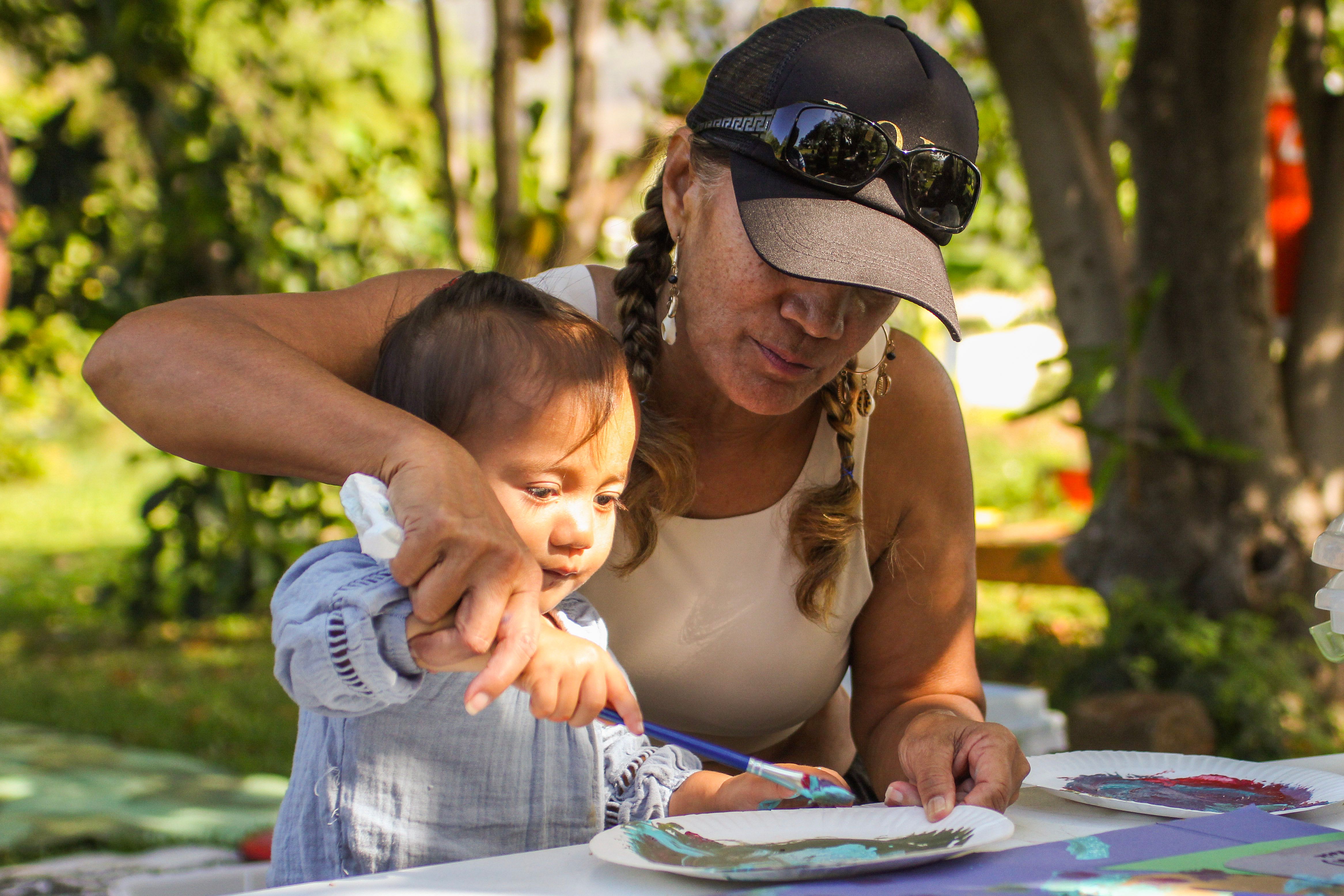 A person wearing a hat guides a child’s hand holding a paintbrush over a piece of paper on a table.