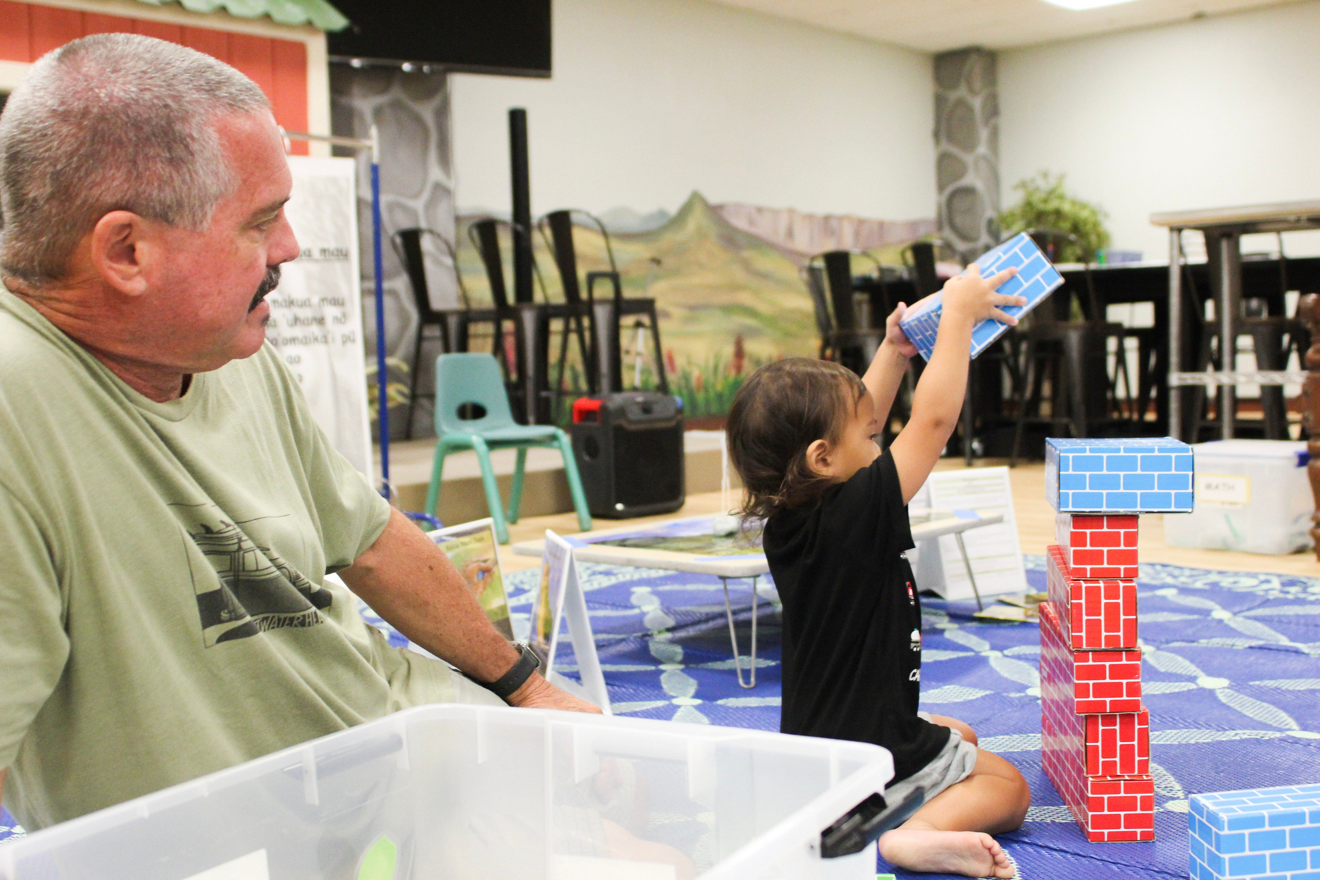 A man with gray hair and a mustache sits on the floor watching a child stack cardboard bricks on top of each other.