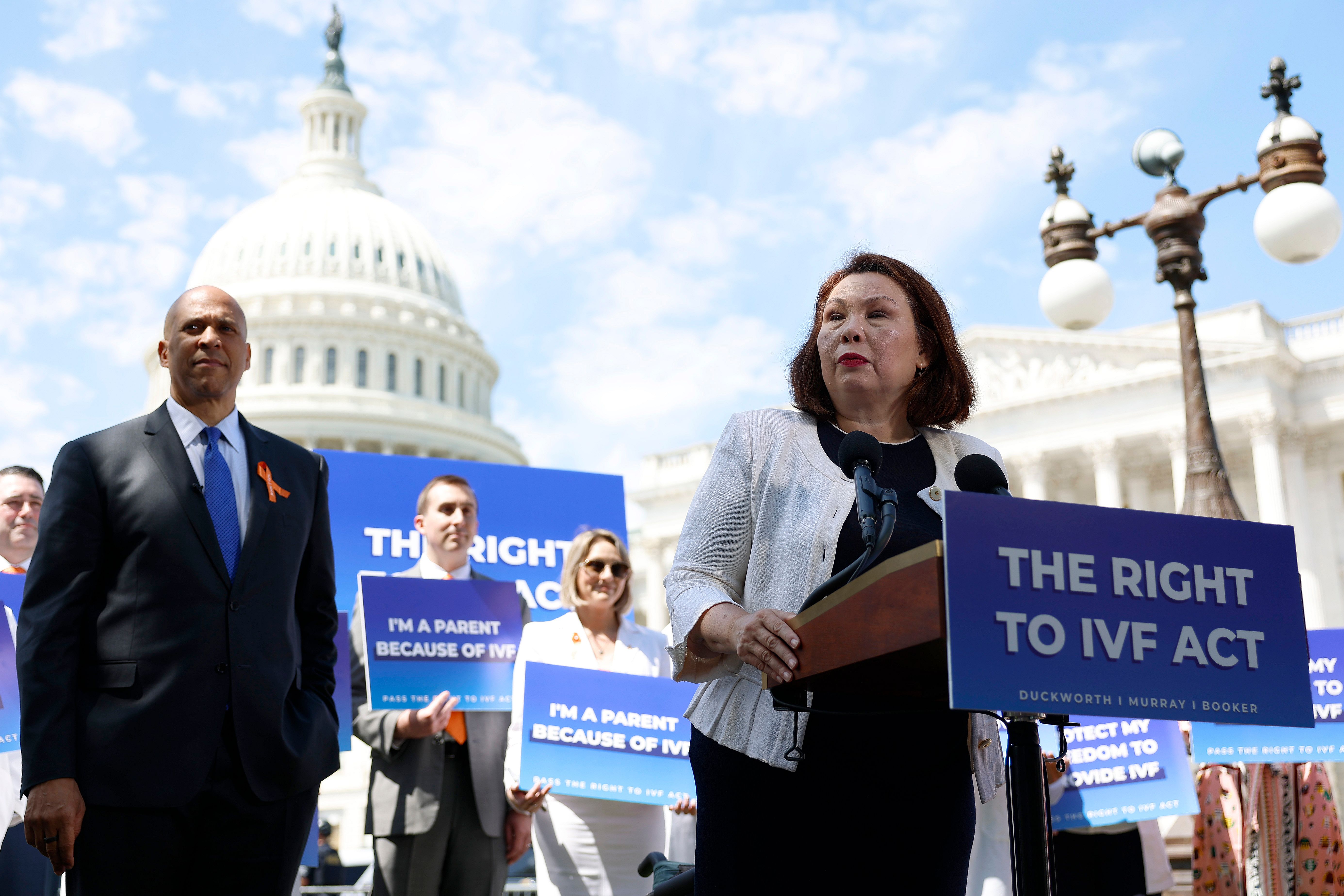 Senator Tammy Duckworth speaks alongside Senator Cory Booker during a news conference on access to IVF treatments outside of the U.S. Capitol Building. The podium in front of her has a poster that reads, The Right to IVF Act while people behind her hold signs that say, I'm a parent because of IVF.