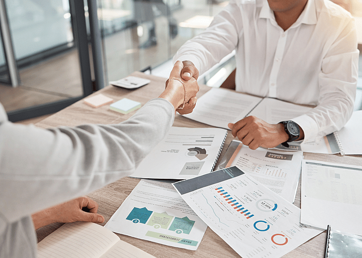 Two people shaking hands over an office desk with charts and reports.