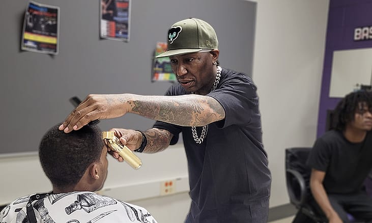A person sits in a chair wearing a plastic cape while a man wearing a cap holds clippers in front of a the person’s forehead.