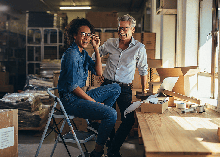 Two people smiling in a small warehouse surrounded by boxes. 