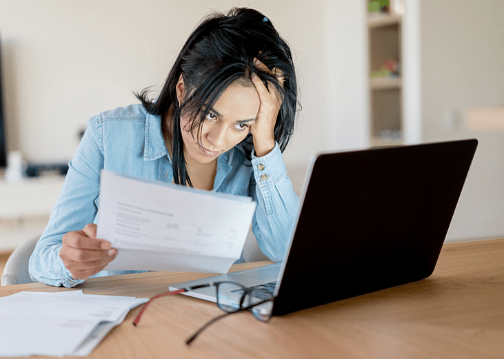 A woman seated at a desk in front of a laptop, holing paperwork and looking frustrated.