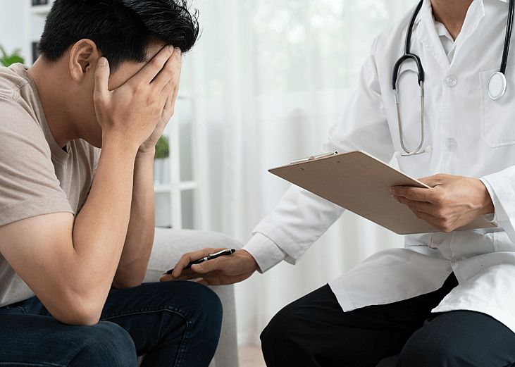 A man sits with his head in his hands, being consulted by a psychologist
