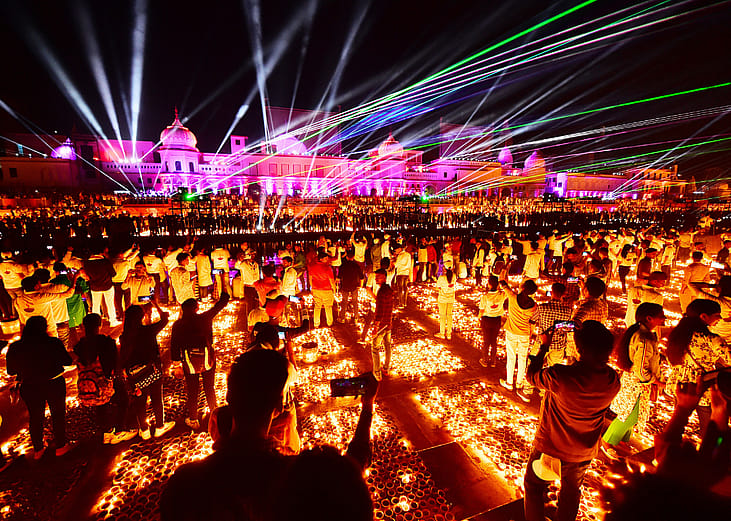A large crowd of people stand in front of a palace with many light beams shining upward, surrounded by candles on the ground.