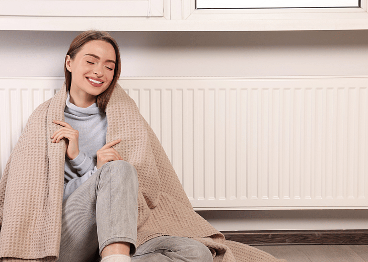 A young woman sits on the floor, wrapped in a blanket, near a wall radiator unit