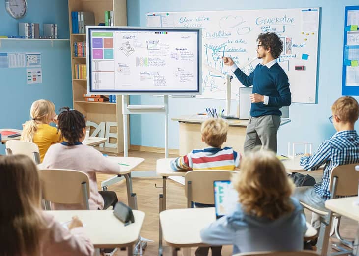 classroom from POV of students watching teacher at a screen in front