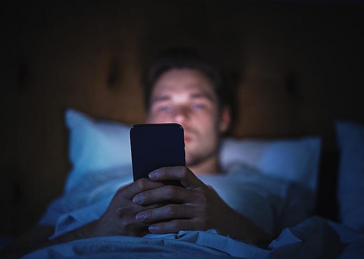 A man browsing on a smartphone while lying in bed at night.