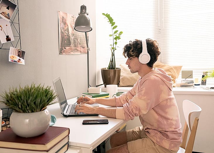 Person wearing headphones and pink tie-dye sweatshirt works peacefully on laptop in room with window light.