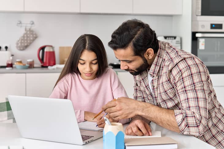 A father and his daughter using a laptop and writing down notes to help with homework.