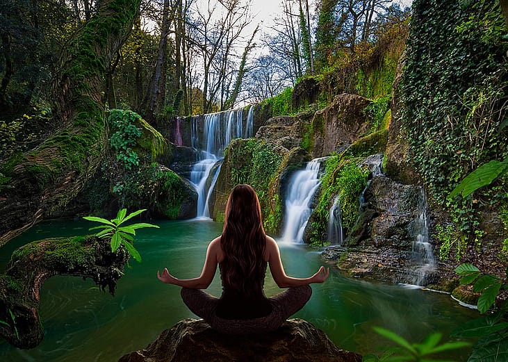 Person sitting in yoga lotus pose facing away from viewer looking at waterfall. 