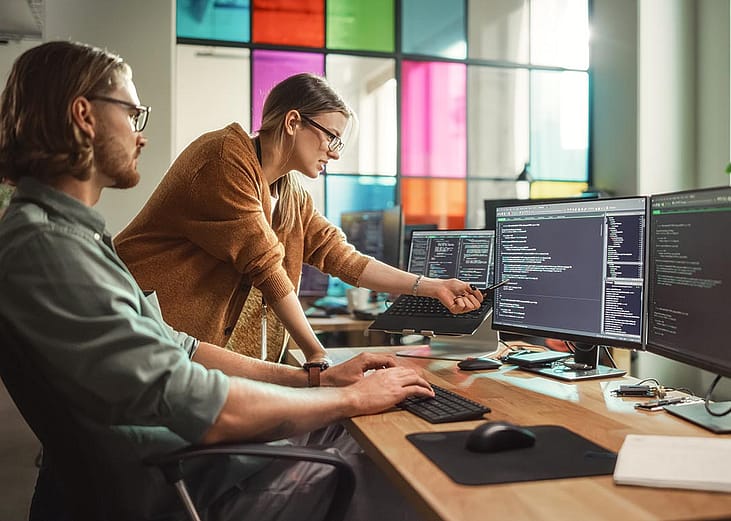 A man working with multiple monitors are engaged in a discussion with his female coworker who points at one of the screens.