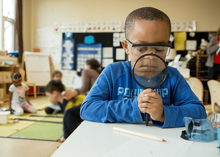A child in a classroom looks through a magnifying glass down at learning materials on his desk. The magnifying glass makes the bottom of his face appear larger.