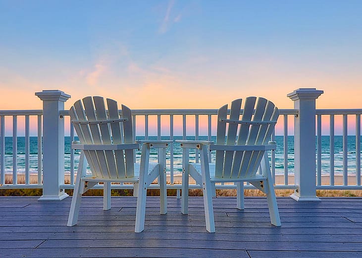 Two coastal style outdoor chairs facing the beac during sunset.