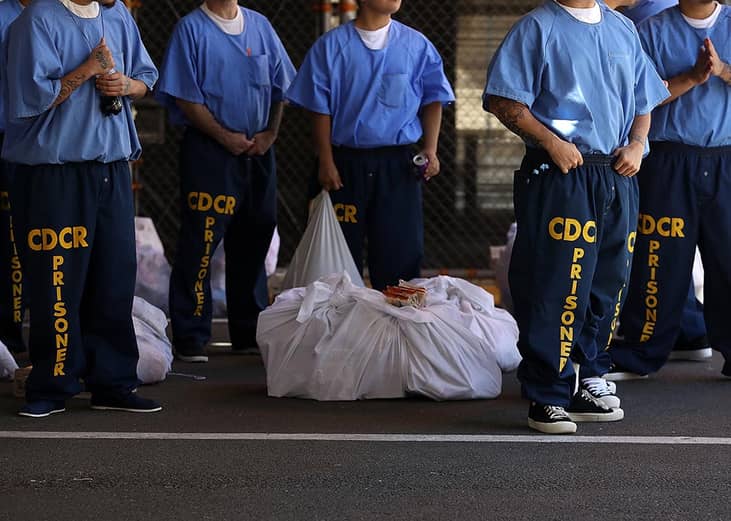 Inmates at San Quentin State Prison shown from the neck down wearing blue uniforms and navy sweat pants that say "CDCR prisoner" wait in line in 2016.