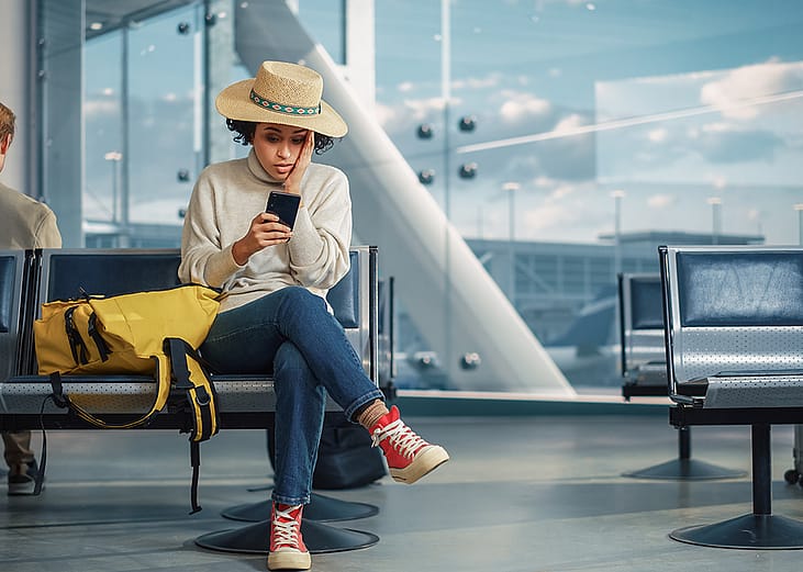 A woman in an airport is checking flight updates in phone while looking worried.