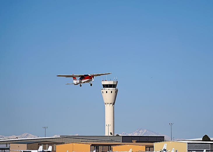 A single engine plane soaring near an airport control tower.