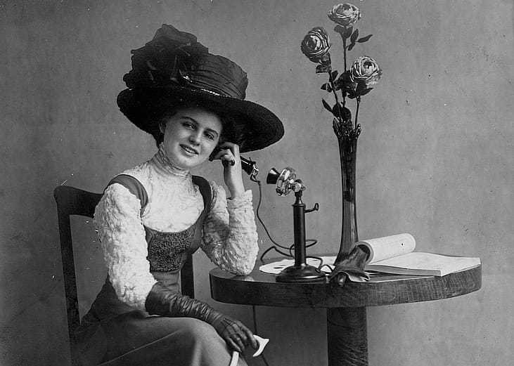 Person with hat seated at table talking on an early standing telephone.