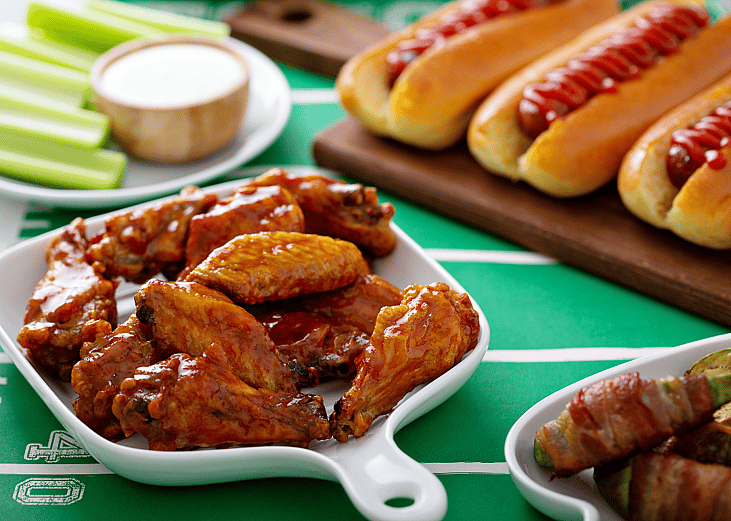 A table with glazed chicken wings, hot dogs, celery sticks and dip