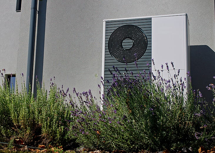 A modern outdoor heat pump mounted on a wall of a home surrounded by lavender plants.