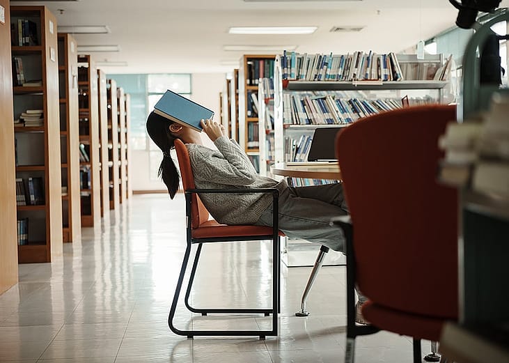 A female college student in a library with an open book over her face seemingly tired of reading.