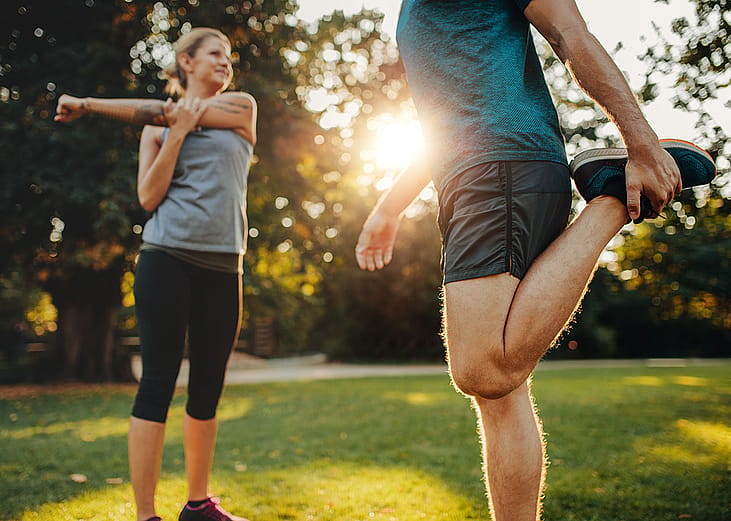A man and a woman both stretching outdoors during a morning warmup.