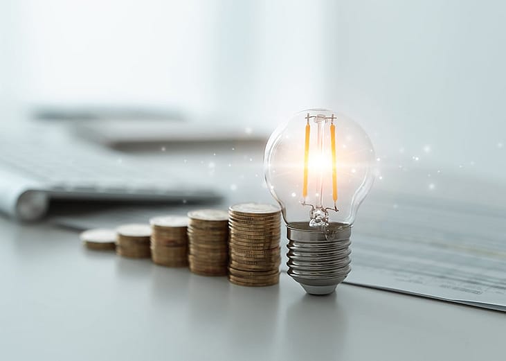 A light bulb and rows of coins decreasing against the background of financial reports as a concept of reducing tax costs.