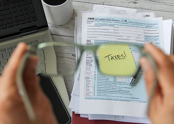 A selective focus of person holding eyeglasses over a tax form and the word "taxes" on a note.