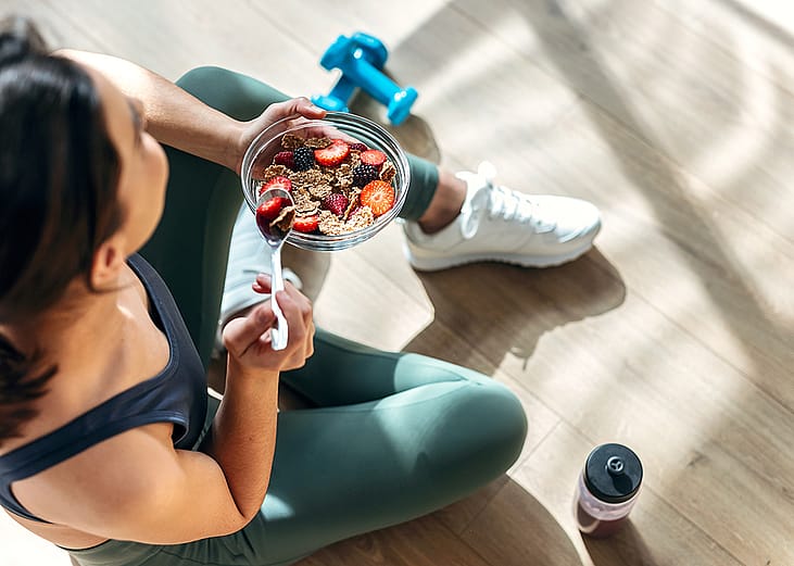 A young woman sitting on the floor post-workout and eating a bowl of oatmeal with fruits.