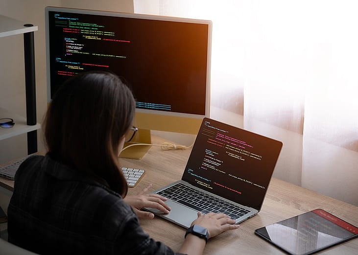 A woman working from home with a laptop and separate monitor equipment showing tech-related interface.