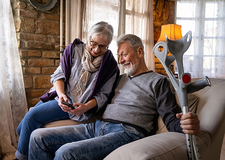 An elder couple where the wife shows her phone to her husband who is holding on to a pair of crutches while sitting on a sofa.