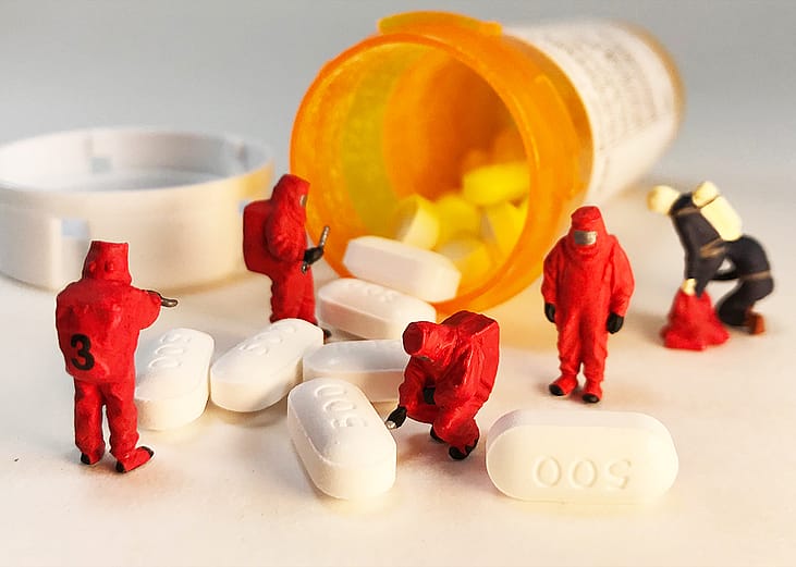 Spilled opioid pills from the bottle and miniature first responders in biohazard suits are investigating it.