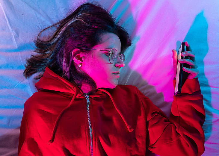 A young girl looking at her smartphone and doom-scrolling on the bed in her neon-lit room.