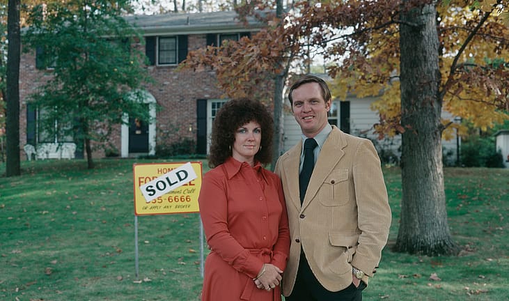 Portrait of couple standing next to For Sale sign in front of house