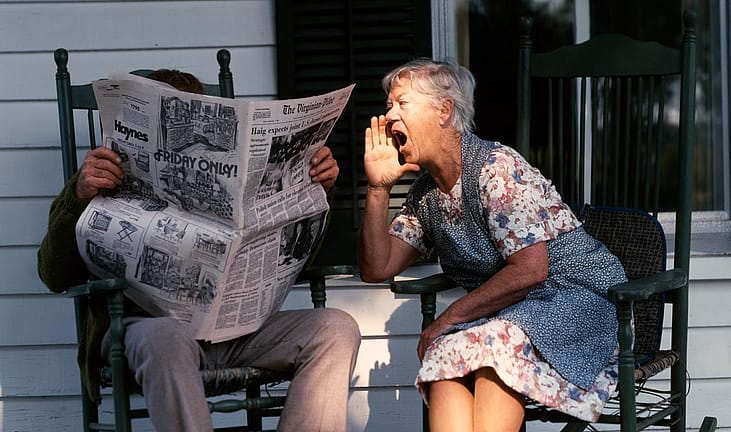 Senior couple sitting on a porch in rocking chairs. Husband is reading the paper and the wife is shouting at him. 