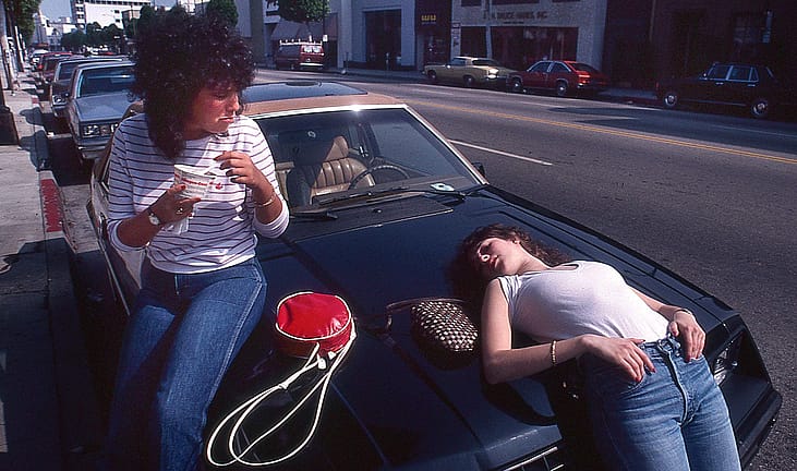 Two teenage girls sit and lie on the hood of a parked car on a Beverly Hills street, one eating from a red thermos cup, 1982.