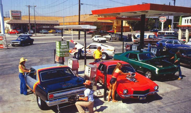 Customized muscle cars, including a red Camaro with a hood-mounted supercharger and a car with flame paint detailing, being fueled at a Texaco gas station with prices around $1.24–$1.30 a gallon.