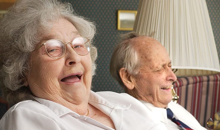 A senior woman with curly gray hair and glasses laughs with her head tilted back, while an older bald man beside her laughs with his eyes closed, both seated in a cozy living room with striped upholstery and a lamp.