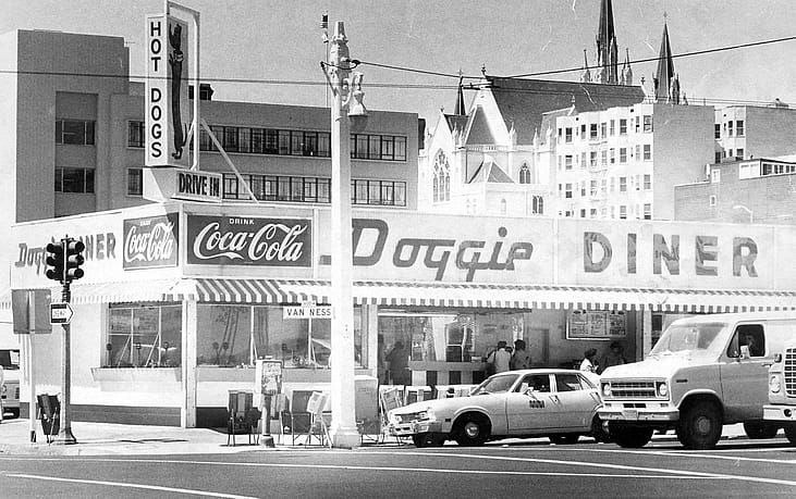 Black-and-white photo of a Doggie Diner storefront with customers at the order window, a giant dachshund hot dog sign, and Coca-Cola signage, cars parked on the street outside.