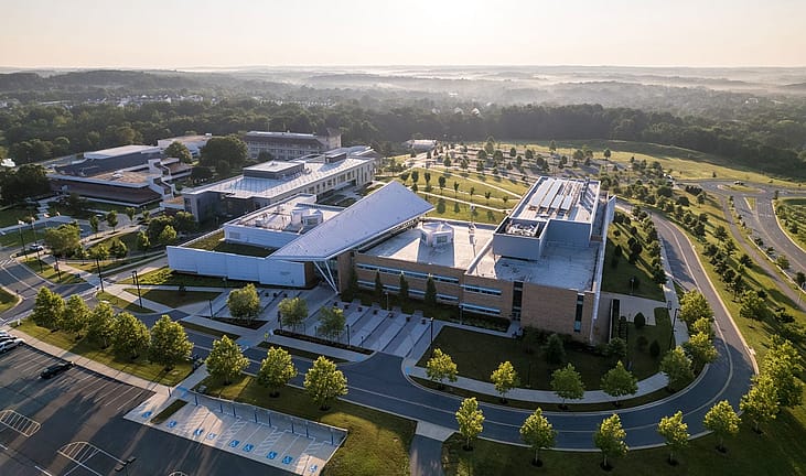 Aerial view of a modern community college campus with interconnected buildings, a landscaped courtyard, and surrounding parking lots at sunrise.