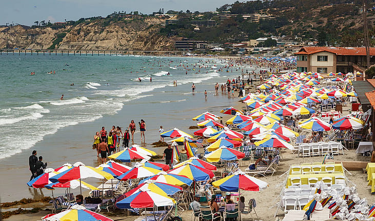 Rows of colorful umbrellas fill a packed beach with swimmers and surfers in the water, capturing a classic summer day at the coast.