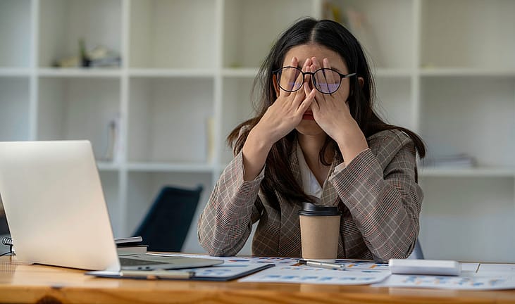 Stressed office worker rubbing her eyes at a desk covered in paperwork.