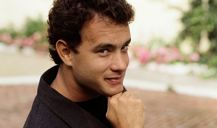 Tom Hanks in the late 1980s, wearing a dark jacket, glancing over his shoulder with a slight smile, blurred pink flowers in the background.