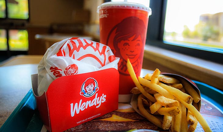 A Wendy's meal tray with a burger in branded wrapper, a Wendy's-logo fry container and drink cup featuring the mascot, on a restaurant table by a window.
